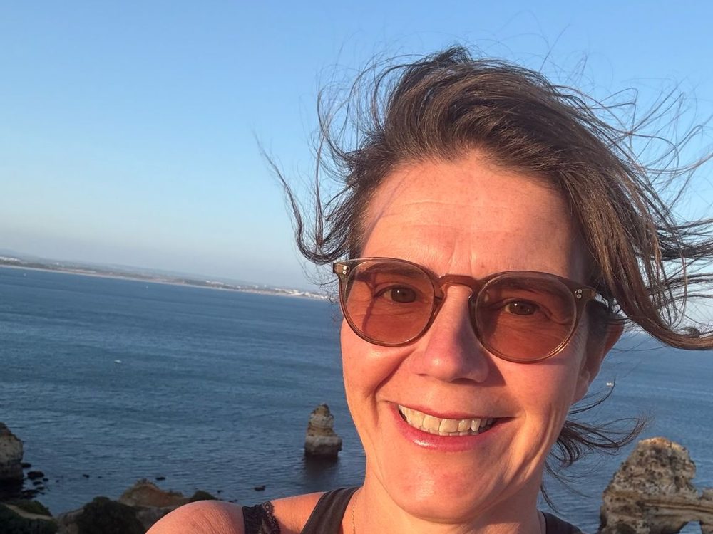 Marlies van Beek, lachend in de wind aan de kust, met de zee en rotsen achter haar